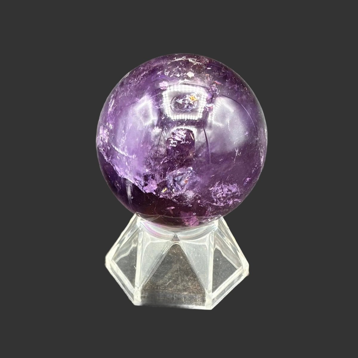 Back side of this polished Amethyst sphere on a clear acrylic stand, in front of a black background.