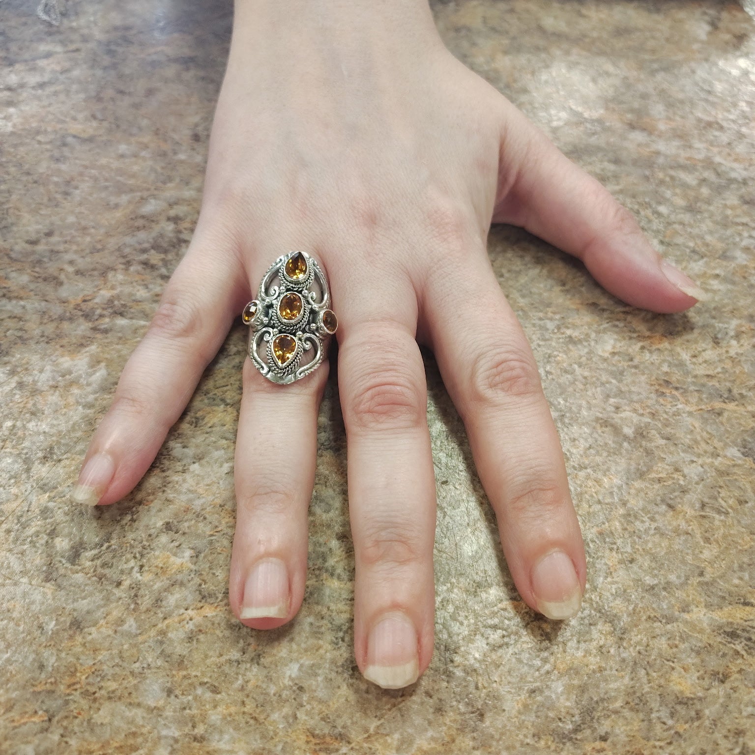 Hand wearing a silver ring with a heart design and yellow gemstone on a marble surface