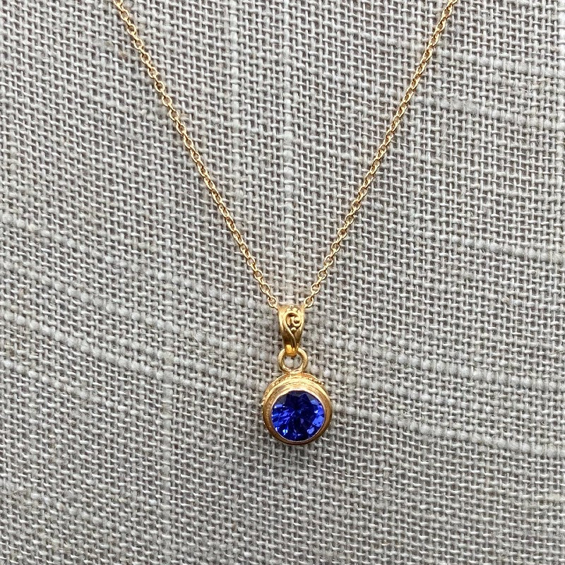 Close Up Of Unique Tanzanite Gemstone Gold Necklace, Gemstone Is Bright Blue And Round. Chain Is Gold
