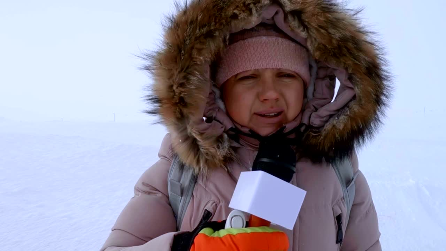 Child in a winter coat with a fur-lined hood holding a microphone outdoors.