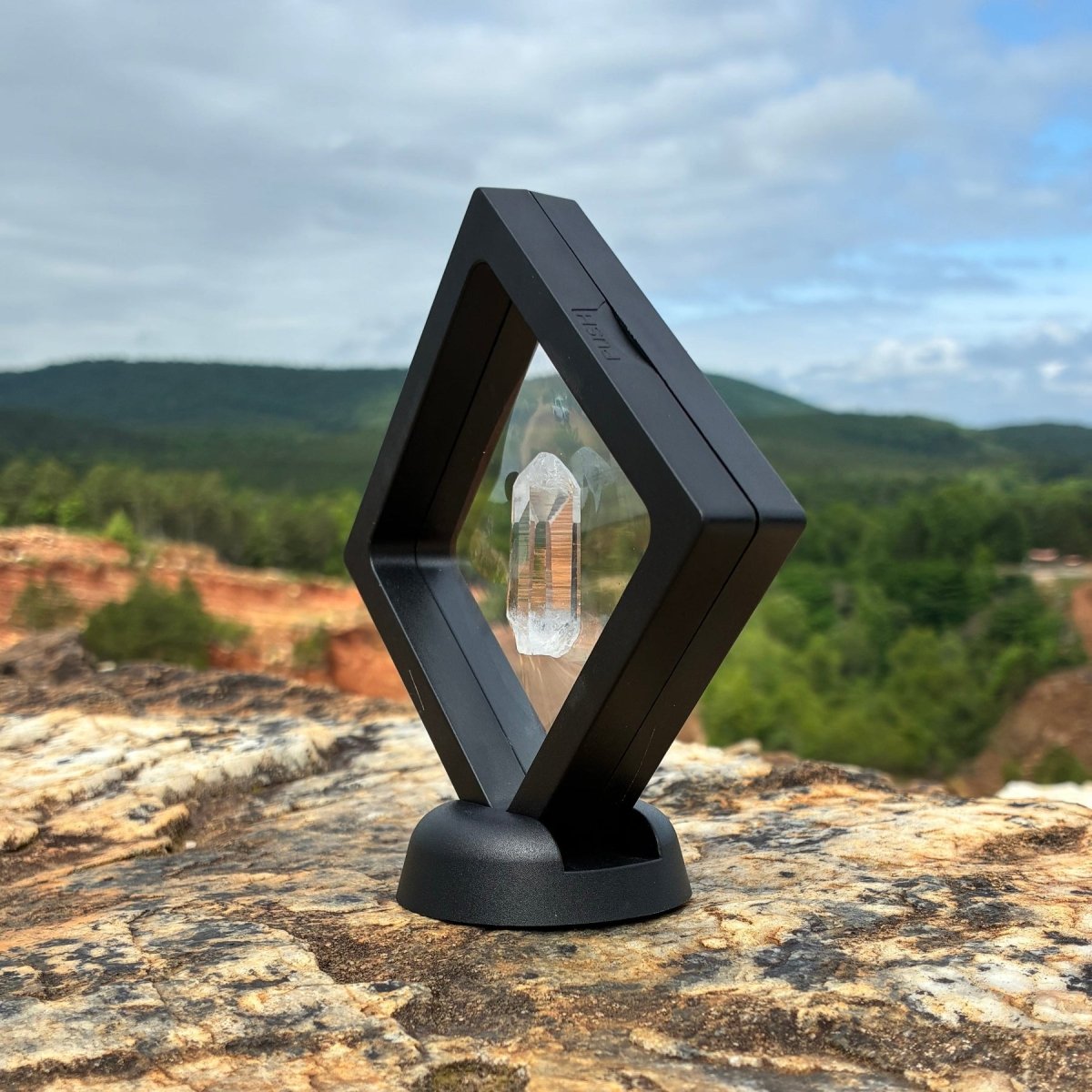 Side view of this water clear quartz crystal point, in a clear frame with a base,and on a boulder outdoors.