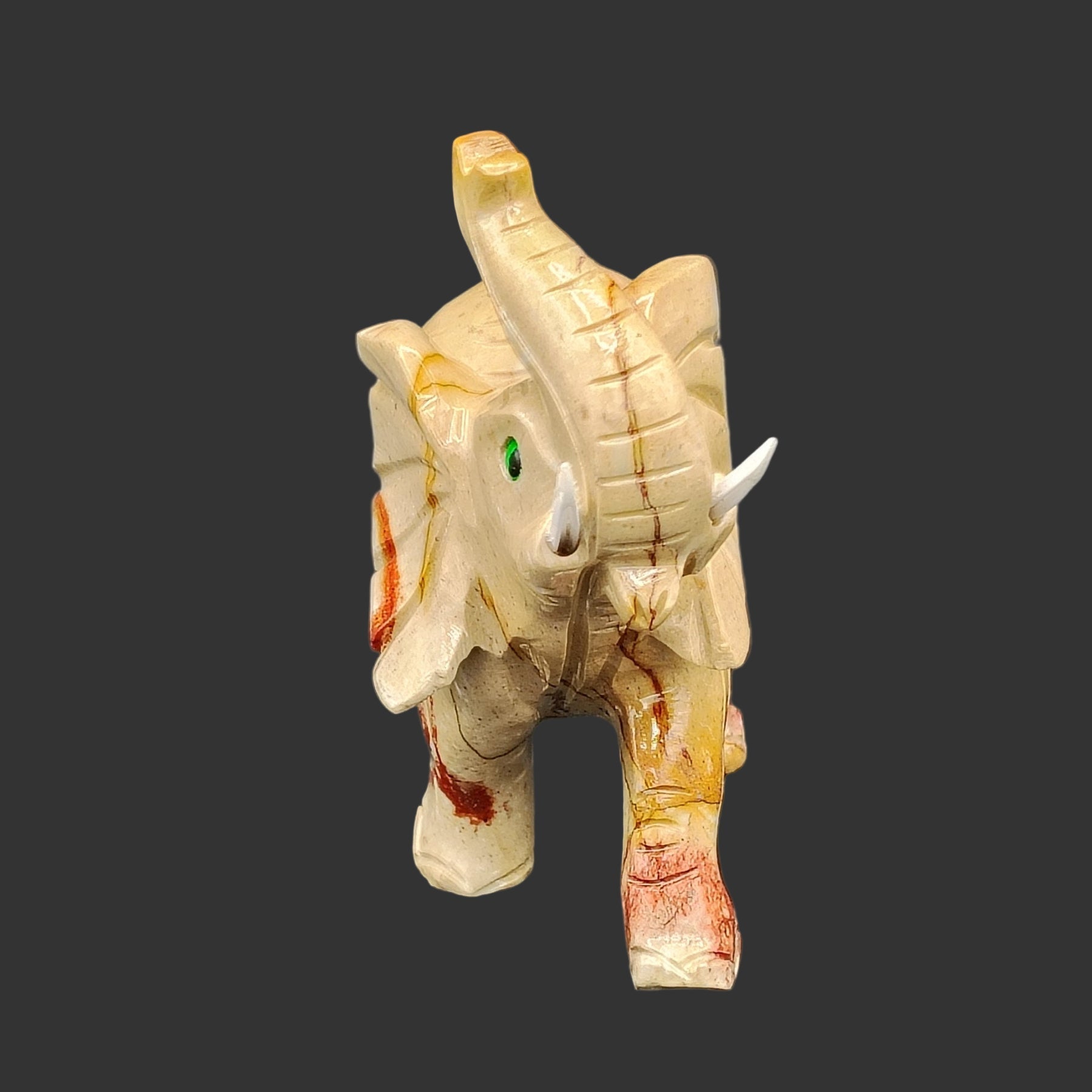 This is the front view of this hand-carved soapstone elephant. It is mostly beige with some red color on his legs, green eyes, and white tusks.